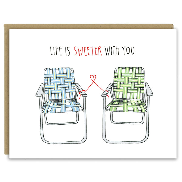 A greeting card showing a hand-drawn illustration of a pair of old-fashioned lawn chairs with webbing and metal frames, one in blues and one in greens. A red thread ties the two chairs together and forms a small heart shape between them. A hand-lettered message reads, "Life is sweeter with you." Shown with a Kraft paper envelope on a white background. 