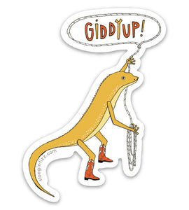 A vinyl sticker of a hand-drawn yellow lizard wearing red cowboy boots, swinging a lasso with its left hand, with the words giddy up hand-lettered inside the lasso, on a white background.
