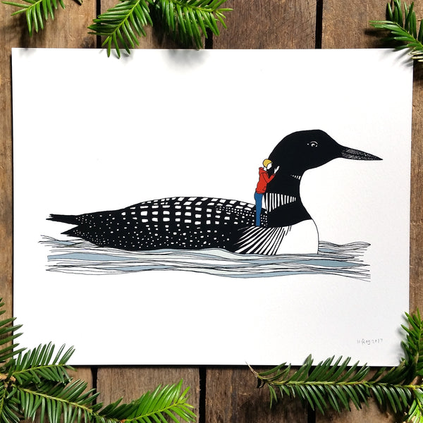 A print of a hand-drawn illustration of a black and white loon floating gently on the waves. A small woman with a yellow bob wearing jeans and a burnt orange hoodie stands on the loon's back with one hand resting on its neck. Shown on a wood background with evergreen branches around it.