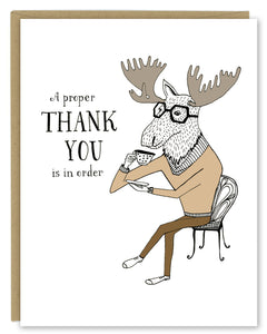 A greeting card showing a hand-drawn illustration of a moose wearing a tan cowl-necked sweater, sitting cross-legged on a wire cafe chair, daintily raising a cup of tea to his mouth. A hand-lettered message reads, "A proper thank you is in order." Shown with a Kraft paper envelope on a white background. 
