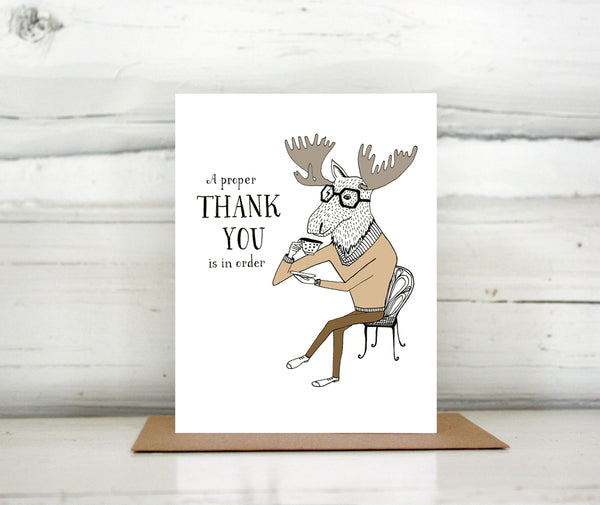 A greeting card showing a hand-drawn illustration of a moose sitting cross-legged on a wire cafe chair, daintily raising a cup of tea to his mouth. A hand-lettered message reads, "A proper thank you is in order." Shown standing on a Kraft paper envelope in front of a white-washed log wall. 