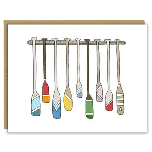 A greeting card with a hand-drawn illustration of a rack full of colorful oars and paddles each decorated with different color and stripes. Shown with a Kraft paper envelope on a white background. 