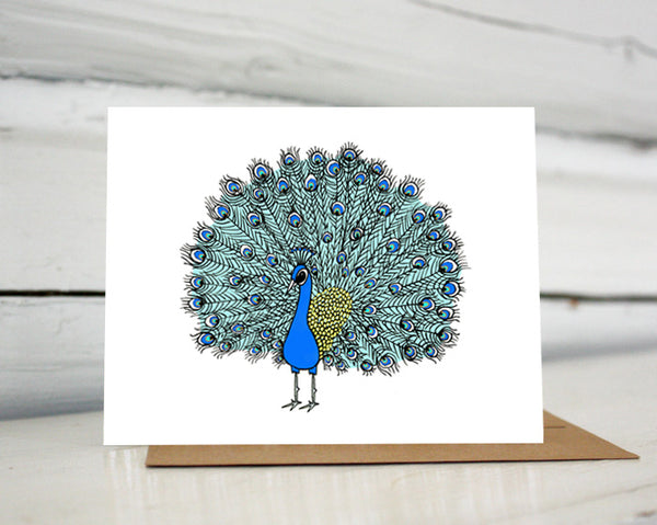 A greeting card with a hand-drawn ink illustration of peacock with its showy tail feathers in full plume. Shown standing on a Kraft paper envelope in front of a white-washed log wall. 