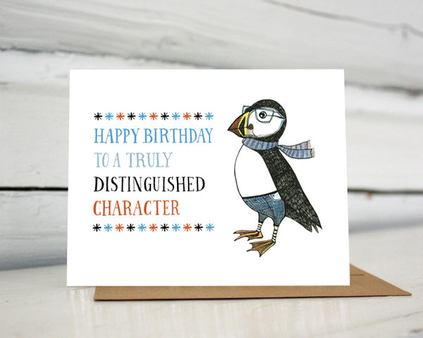 A greeting card with a hand-drawn ink illustration of a dapper puffin wearing glasses, jeans and a scarf. A hand-lettered message reads, "Happy Birthday to a truly distinguished character." Shown standing on a Kraft paper envelope in front of a white-washed log wall. 