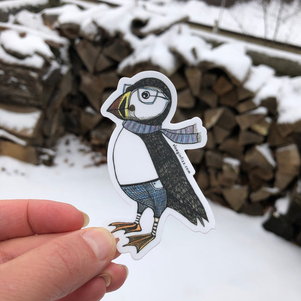 A hand holding up a sticker with a hand-drawn ink illustration of a dapper puffin in glasses, jeans and a scarf in front of a snowy background.