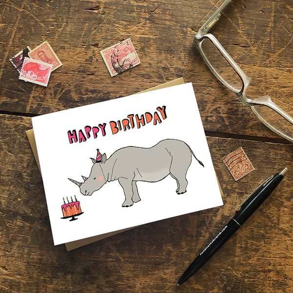 A greeting card showing a hand-drawn illustration of a rhinoceros wearing a pink and black striped party hat that matches stripes on his horns, looking at an orange and pink birthday cake. A hand-lettered message reads, "Happy Birthday." Shown with a Kraft paper envelope on a worn wooden surface with reading glasses, stamps and a pen. 