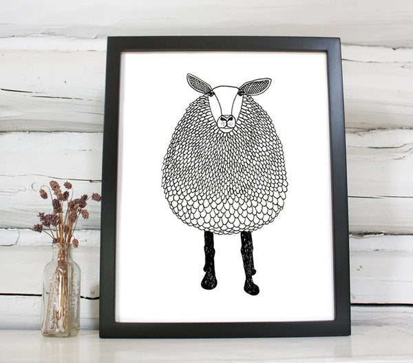 A print of a hand-drawn black and white ink illustration of a sheep. Shown in a black frame on a shelf with a small vase of dried flowers next to it, in front of a whitewashed log wall. 