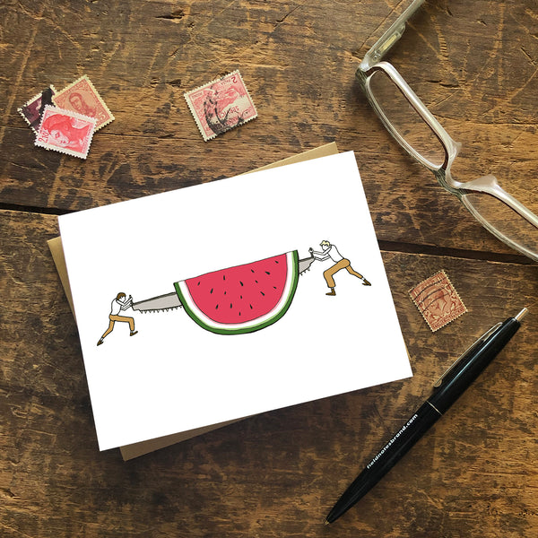 A greeting card shows a pair of loggers sawing off a slice of a giant watermelon in a hand-drawn illustration. Seen with a Kraft paper envelope on a wood surface with stamps, a pen and reading glasses. 